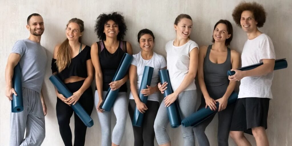 imgi_32_Group_portrait_of_happy_diverse_people_with_yoga_mats_in_hands_posing_for_photo_at_grey_wall_background_looking_at_camera_smil